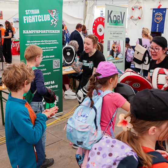 Eine Frau mit Tätowierungen zeigt Kindern Boxhandschuhe an einem Stand mit einem Schild, das Styrian Fightclub sagt. Andere Leute unterhalten sich mit der Frau.