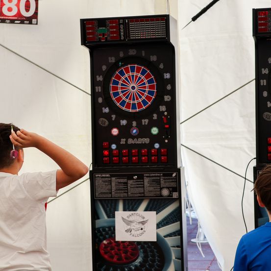 Ein junger Junge wirft Darts auf einen Arcade-Dartautomaten, gekennzeichnet mit Dartclub Falcons. Ein anderer Junge beobachtet von hinten.