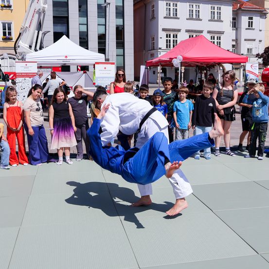 Zwei Personen üben Kampfsport in einem blauen Anzug, während Kinder auf der Straße zusehen. Ein Zelt und Gebäude sind im Hintergrund.
