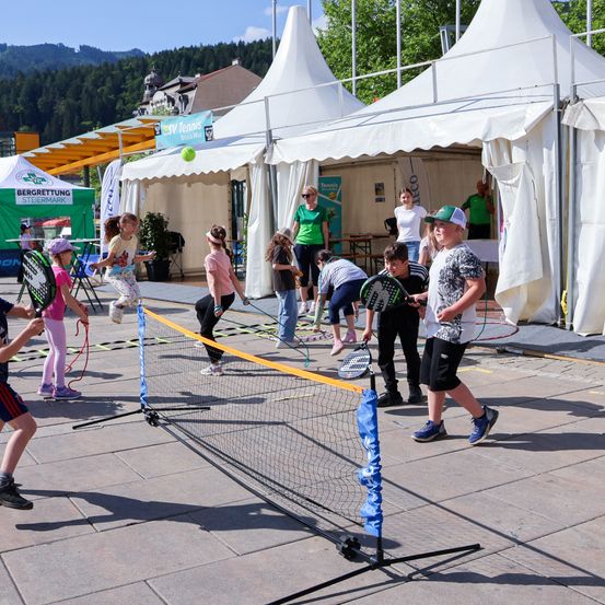 Kinder spielen Tennis auf einem Freiluftplatz mit Zelten im Hintergrund. Sie schlagen einen Ball über ein Netz und einige Erwachsene beobachten.