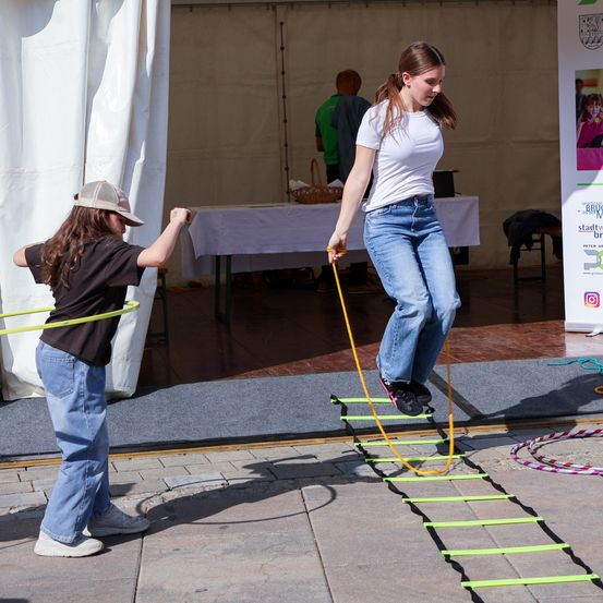 Ein Mädchen versucht, auf einer gelben Leiter zu balancieren, während ein anderes Mädchen ein Hula-Hoop hält. Ein Tisch und ein Mann sind im Hintergrund.