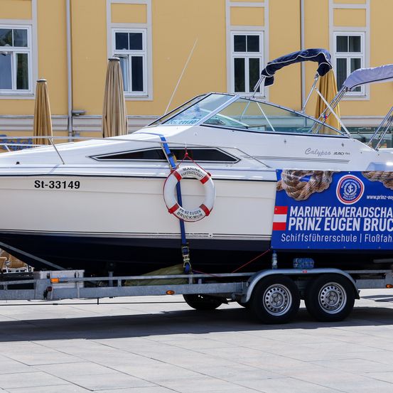 Ein weißes Boot mit der Nummer St-33149 steht auf einem Anhänger vor einem gelben Gebäude. Am Boot ist ein Rettungsring befestigt.