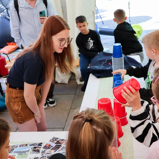 Eine Frau und Kinder nehmen an einem Becher-Stapelspiel teil. Die Frau gibt Anweisungen, während ein Kind einen roten Becher hält. Andere Kinder und Erwachsene beobachten.