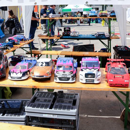 Eine Ausstellung von ferngesteuerten Autos ist unter einem Zelt aufgebaut. Verschiedene Autos in verschiedenen Farben stehen auf dem Tisch. Eine Frau und ein Mann stehen hinter dem Tisch.