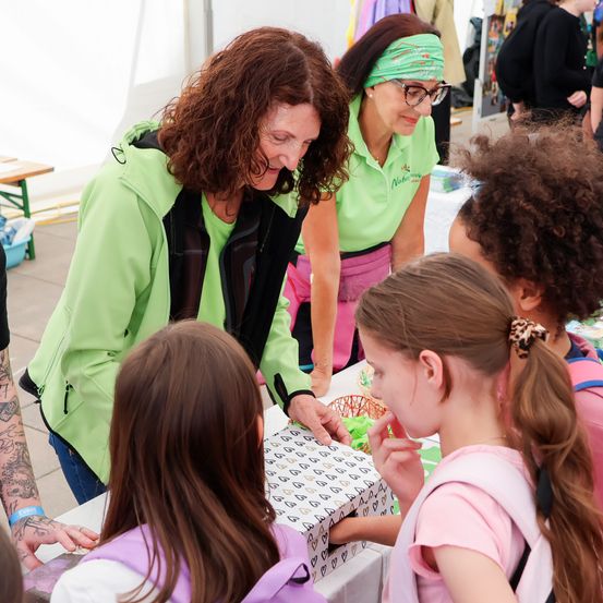 Eine Frau in einer grünen Jacke lächelt und verteilt Gegenstände an Kinder an einem Tisch.