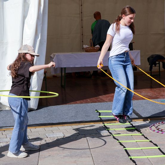 Zwei Mädchen spielen mit einem Hula-Hoop und einer Leiter. Eine hält den Hula-Hoop, während die andere die Leiter erklimmt. Sie befinden sich in einem Zelt.