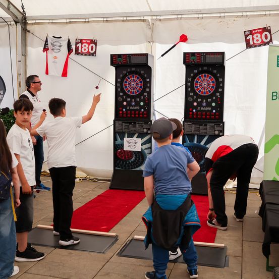 Eine Gruppe von Kindern und Erwachsenen spielt Darts in einem Zelt mit rotem Teppich und zwei Dart-Boards. Die Punktestände von 180 sind an der Wand angebracht.
