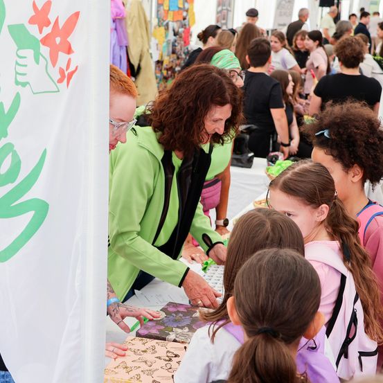 Eine Menschenmenge bei einer Veranstaltung, eine Frau in Grün hilft Kindern bei der Betrachtung von Schmetterlingsdesigns auf einem Tisch.