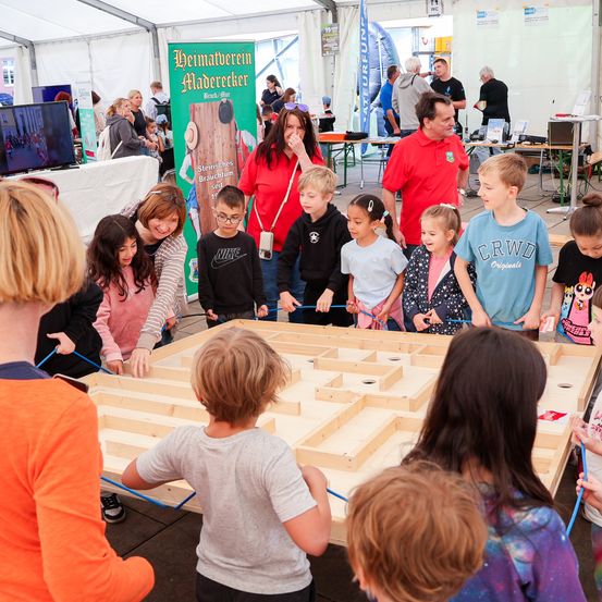 Kinder und Erwachsene sind bei einem Holzlabyrinth-Spiel in einem Zelt-Event versammelt, einige lächeln, andere konzentrieren sich auf das Spiel.