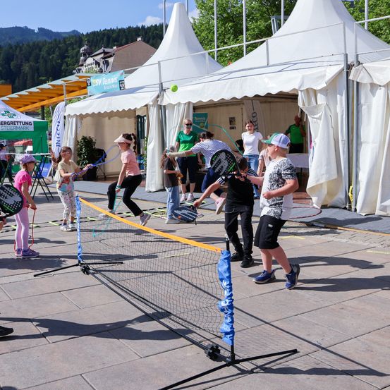 Eine Gruppe von Kindern und Erwachsenen spielt Tennis auf einem Freiluftplatz. Vor ihnen ist ein Netz aufgestellt. Hinter ihnen befinden sich weiße Zelte mit einem Banner.