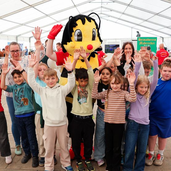 Kinder und Erwachsene stehen in einer Gruppe und winken mit den Händen. Ein großes Bienchen-Maskottchen steht unter ihnen. Einige Leute tragen eine Brille, und eine Frau lächelt in die Kamera. Ein grüner Banner und ein weißes Zelt sind im Hintergrund.