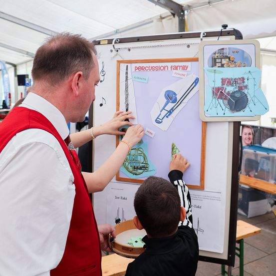 Ein Erwachsener in einer roten Weste bringt einem jungen Jungen bei, ein Perkussionsinstrument zu spielen, bei einer Veranstaltung. Das Kind hält eine kleine Trommel, während der Erwachsene auf ein Poster mit Musikinstrumenten zeigt.