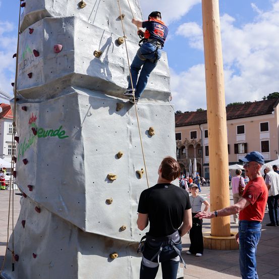 Eine Person klettert an einer großen Kletterwand, während eine andere zusieht. Sie sind im Freien in der Nähe eines Gebäudes mit mehreren Menschen im Hintergrund.