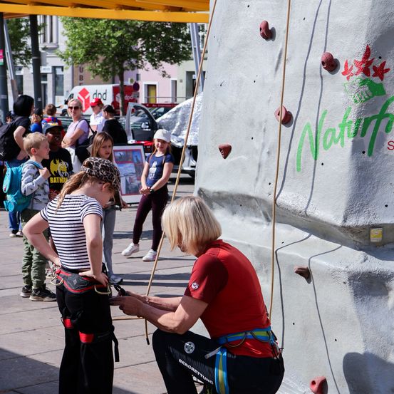 Eine erwachsene Frau hilft einem jungen Madchen, ihr Klettergurt zu befestigen, an einer Kletterwand, die von einer Menschenmenge beobachtet wird.