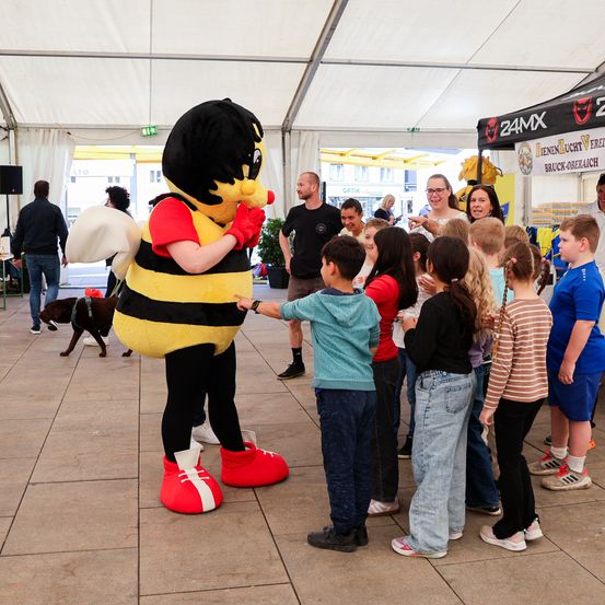 Eine als Biene verkleidete Maskottchen interagiert mit einer Gruppe von Kindern, während Erwachsene zuschauen. Die Szene findet unter einem Zelt mit einem Zelt im Hintergrund statt.