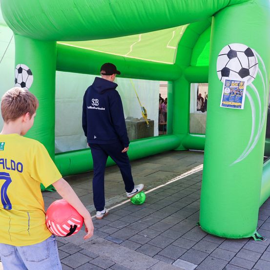 Zwei Kinder spielen mit Fußballbällen in einem grünen aufblasbaren Fußballtor. Ein Mann in einer blauen Kapuzenjacke steht neben dem Tor und hält einen grünen Ball.