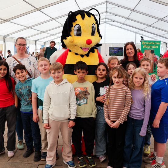 Eine Gruppe von Kindern und zwei Erwachsenen posiert mit einem riesigen Bienenmascot für ein Foto bei einer Veranstaltung im Freien unter einem Zelt.