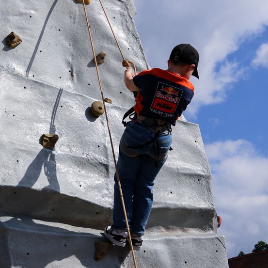 Ein Kind in einem Red Bull-Shirt klettert an einer Indoor-Kletterwand und hält mit beiden Händen ein Seil fest.
