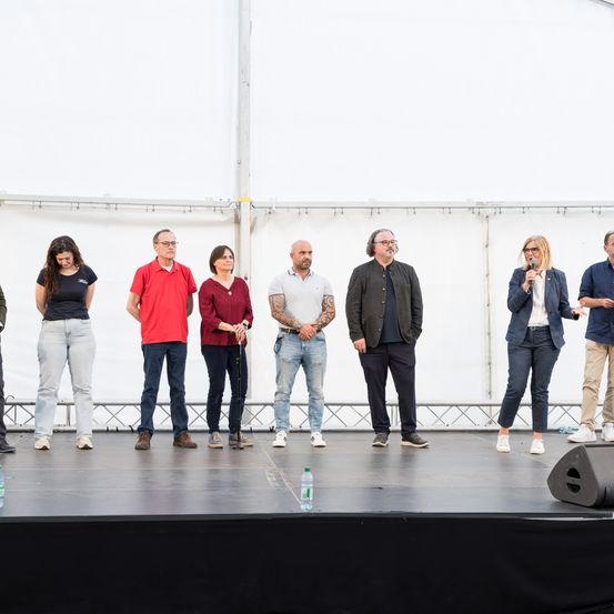 Eine Gruppe von Personen steht mit Mikrofonen auf einer Bühne. Sie scheinen eine Präsentation zu halten. Die Bühne hat einen weißen Hintergrund und ist schwach beleuchtet.