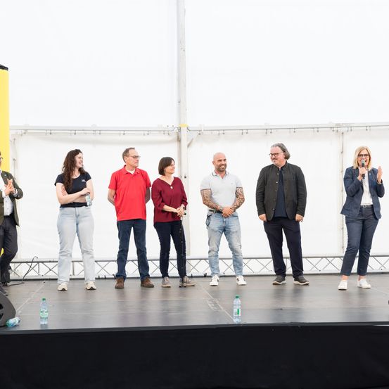 Eine Gruppe von Menschen steht auf der Bühne vor einer weißen Kulisse, in lässiger Kleidung. Sie scheinen sich auf eine Aufführung vorzubereiten.
