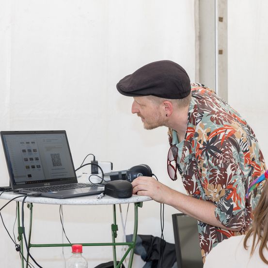 Ein Mann mit Hut arbeitet an einem Laptop an einem Tisch, vor ihm ein Monitor. Er hält eine Maus und scheint etwas zu scannen.