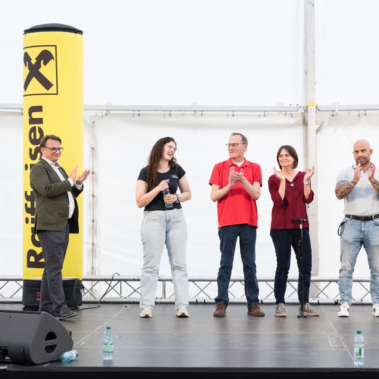 Fünf Personen auf einer Bühne, die Hände klatschen. Ein gelbes Schild mit dem Wort 'Roelfsen' und einem 'X' hinter ihnen. Ein Lautsprecher links und ein Dreibeinstativ rechts.