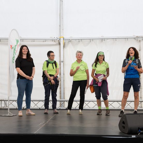 Fünf Frauen stehen auf einer Bühne, möglicherweise bei einer Veranstaltung, und tragen grüne T-Shirts. Eine Frau hat einen Rucksack, und eine andere hält ein Blatt Papier. Ein Lautsprecher steht auf der Bühne, und eine weiße Flagge ist auf der linken Seite.