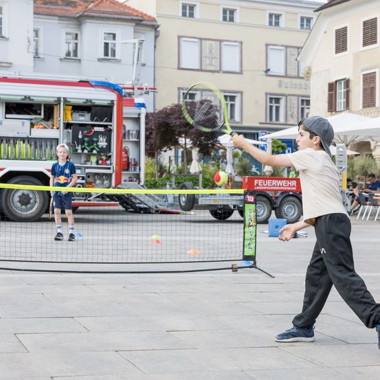 Ein Junge spielt Tennis mit einem Freund auf einem öffentlichen Platz. Ein Feuerwehrwagen steht hinter ihnen.