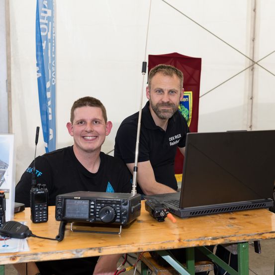 Zwei Männer sitzen an einem Tisch mit Laptop und Funkgeräten. Ein Mann hat ein Funkgerät auf der Schulter. Dahinter befindet sich eine blaue Flagge und ein roter Banner.