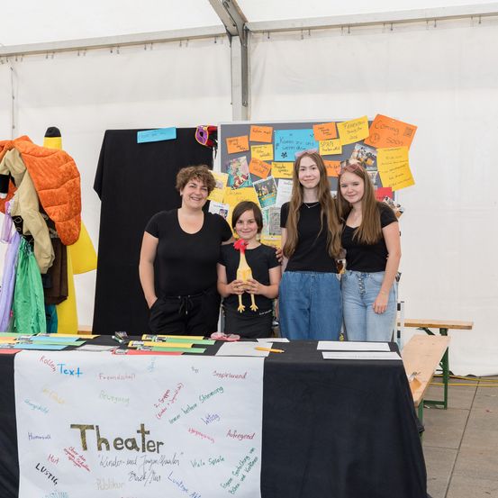 Vier Frauen stehen hinter einem Tisch mit einem Schild, auf dem Theater steht. Ein Mädchen hält eine Puppe in den Händen. Sie lächeln und posieren für ein Foto. Dahinter sind bunte Haftnotizen und eine Tafel an einer weißen Wand befestigt.