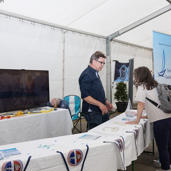 Ein Mann und eine Frau stehen an einem Tisch unter einem Zelt, möglicherweise bei einer Segelveranstaltung. Der Tisch hat eine weiße Tischdecke und verschiedene Gegenstände darauf. Ein großer Fernseher ist an der Wand hinter ihnen montiert.