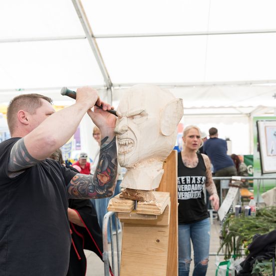 Ein Mann schnitzert eine hölzerne Kopfskulptur, wahrscheinlich bei einer Veranstaltung im Freien. Eine Frau beobachtet ihn, und andere sind im Hintergrund.