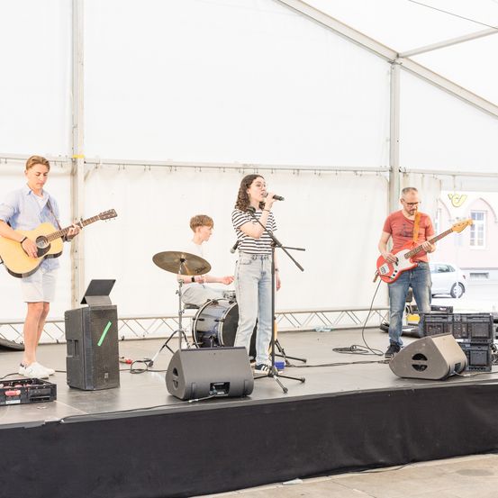 Eine Band tritt auf einer Bühne mit Lautsprechern, Mikrofon und Musikinstrumenten auf. Die Sängerin steht mit einem Mikrofon zwischen zwei Gitarristen. Ein Schlagzeuger spielt hinter der Sängerin.