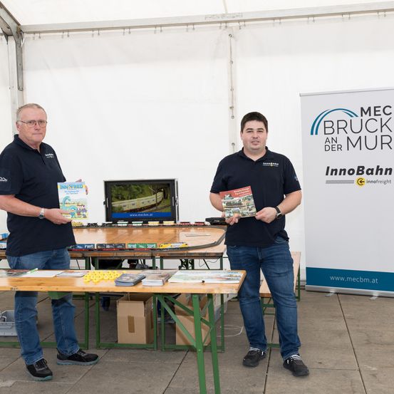 Zwei Männer in schwarzen Poloshirts und Jeans stehen vor einem Tisch mit Broschüren, einem Monitor und einem Modelleisenbahn-Set. Das Banner hinter ihnen sagt MEC BRUCK AN DER MUR InnoBahn.