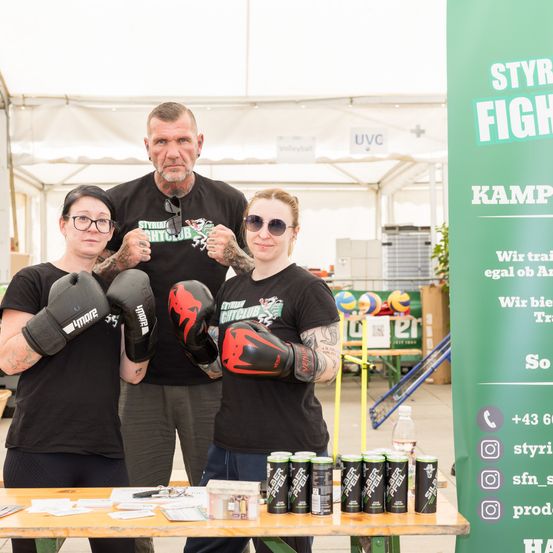 Zwei Frauen in schwarzen Boxhandschuhen posieren für ein Foto mit einem Mann. Sie stehen in einem Zelt mit einem Tisch und Getränken. Ein grüner Banner befindet sich in der Nähe.