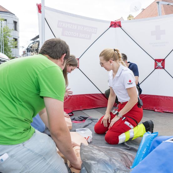 Eine Frau und ein Mann üben auf einer Puppe in einem Zelt, mit einem Roten Kreuz im Hintergrund.