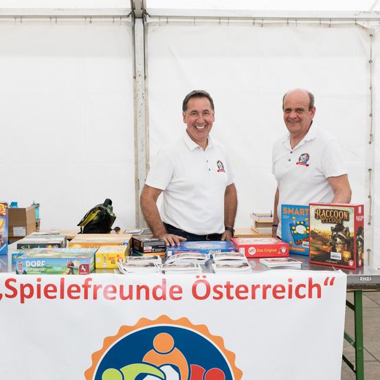 Zwei Männer stehen hinter einem Tisch voller Brettspiele und lächeln in die Kamera. Ein Banner steht 'Spielefreunde Österreich'. Verschiedene Brettspiele sind sichtbar.