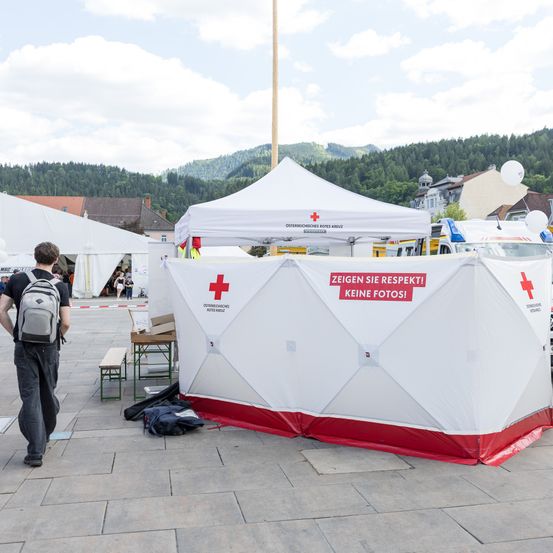 Ein Mann geht an einem weißen Zelt mit rotem Kreuz vorbei. Hinter ihm steht ein weißer Krankenwagen. Das Zelt sagt: 'Zeigen Sie Respekt! Keine Fotos!' Im Hintergrund befinden sich Berge und Gebäude.