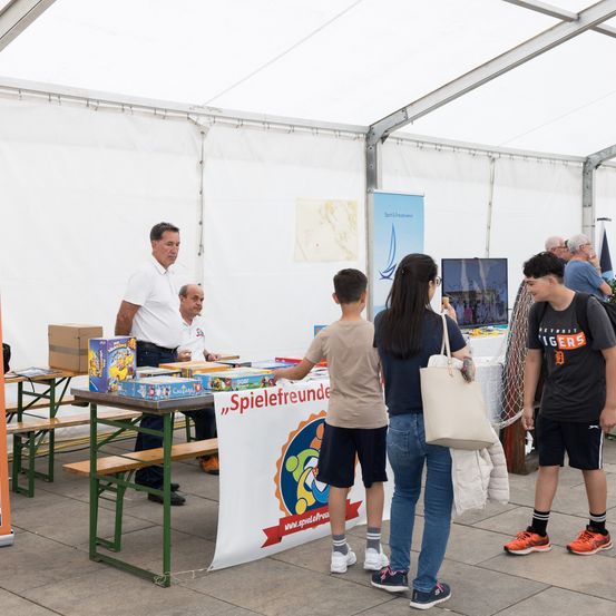 In einem Zelt steht ein Mann hinter einem Tisch mit verschiedenen Brettspielen und interagiert mit einem Jungen und einer Frau, während ein anderer Junge zuschaut.