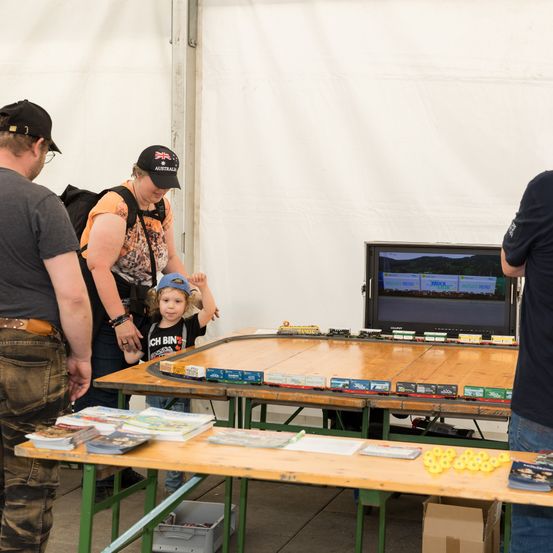 Mehrere Personen stehen um einen Tisch mit einem Spielzeug-Zugset, wobei ein Kind einen Hut trägt. Ein Monitor steht auf dem Tisch, und es gibt Bücher und andere Gegenstände auf dem Tisch und Boden.