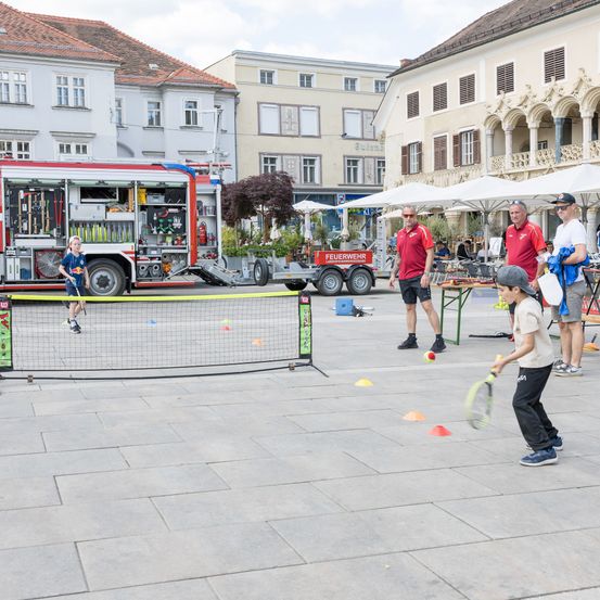 Eine Gruppe von Kindern und Erwachsenen spielt Tennis auf einem gepflasterten Bereich mit einem Netz. Ein Feuerwehrauto ist in der Nähe geparkt, und mehrere Personen stehen herum und beobachten.