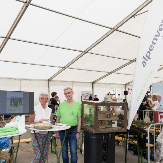 Ein Mann und eine Frau stehen neben einem Tisch unter einem Zelt, ein käfigartiges Miniaturhaus ist ausgestellt.