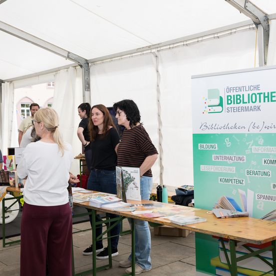 Mehrere Personen sind unter einem weißen Zelt versammelt und stöbern auf einem Tisch durch Bücher. Ein grünes Banner mit dem Text 'Öffentliche Bibliothek' befindet sich hinter ihnen.
