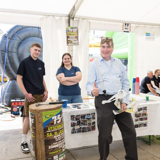 Drei Personen stehen vor einem Stand. Ein Mann gibt einen Daumen hoch. Dahinter steht eine große aufblasbare Struktur und ein Tisch mit Flugblättern.