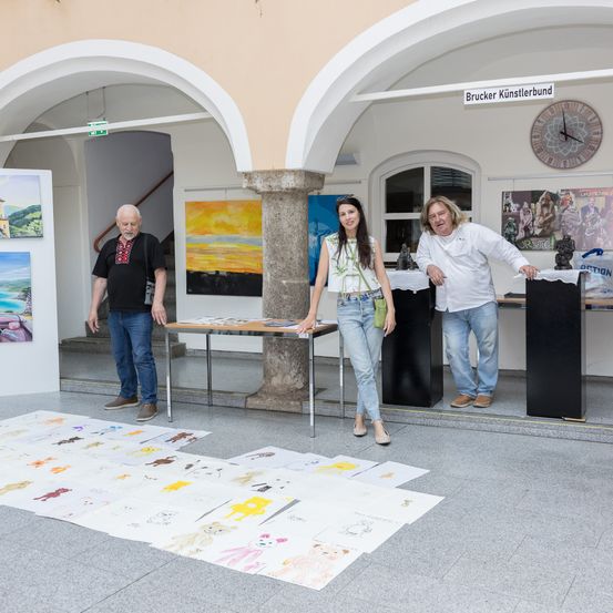 Drei Menschen stehen vor einer Kunstausstellung mit Gemälden und Skulpturen. Sie betrachten Zeichnungen auf dem Boden.