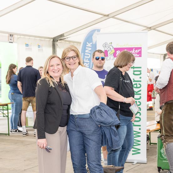 Drei Frauen stehen bei einer Veranstaltung zusammen und lächeln, mit einem Banner im Hintergrund. Dahinter tummeln sich einige Leute, die auf das Banner schauen.