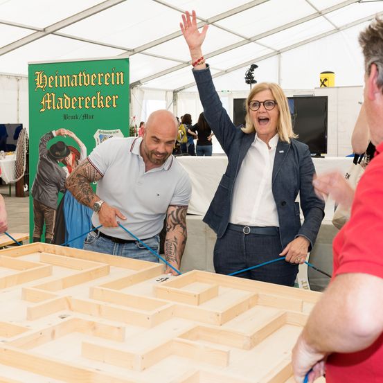 Eine Frau mit einem Ring am Finger hebt die Hand, während zwei Männer lächeln. Sie stehen in der Nähe eines Holzlabyrinths. Ein grüner Banner ist hinter ihnen.