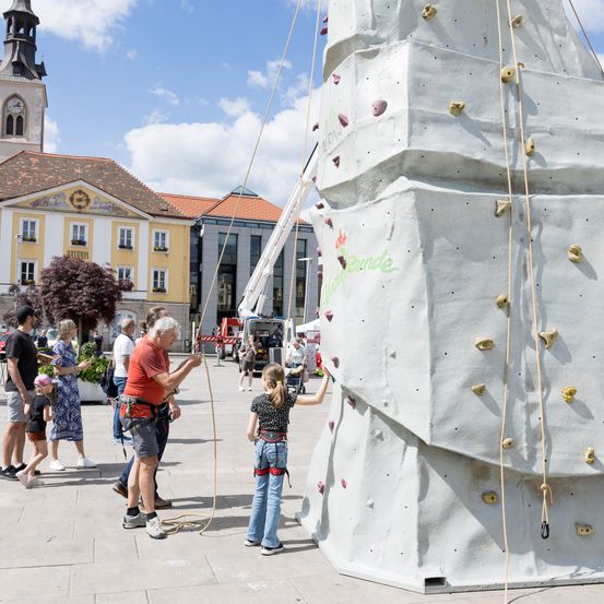 Ein Mann hilft einem jungen Mädchen, eine große Kletterwand in einem Stadtplatz zu erklimmen. Andere Menschen und Gebäude sind im Hintergrund zu sehen.