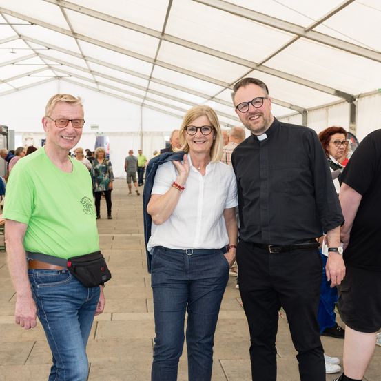 Drei Menschen stehen lächelnd in einem Zelt. Der Mann auf der linken Seite trägt ein grünes Shirt und der Mann auf der rechten Seite trägt ein schwarzes Shirt und eine Brille. Die Frau in der Mitte hält eine blaue Jacke.