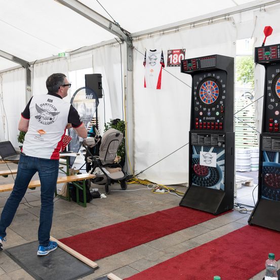 Ein Mann mit einem Shirt mit dem Wort Dartclub wirft einen Dart an einem Arcade-Spiel unter einem weißen Zelt.